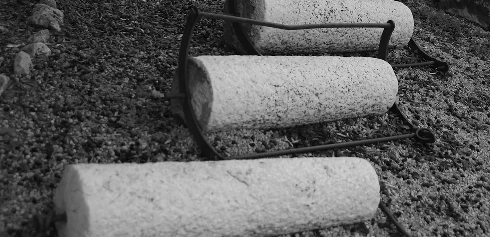 "Black and white image of three stone cylinders joined by a metal frame, placed on a surface of earth and stones. This composition suggests an old agricultural or traditional press equipment, evoking a rustic and historical feel."